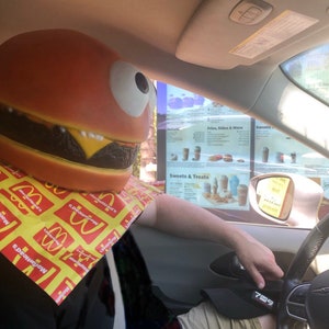 May include: A person wearing a large, red and brown hamburger costume with a yellow McDonald's napkin around their neck, sitting in a car at a drive-thru window. The menu board for the drive-thru is visible in the background.
