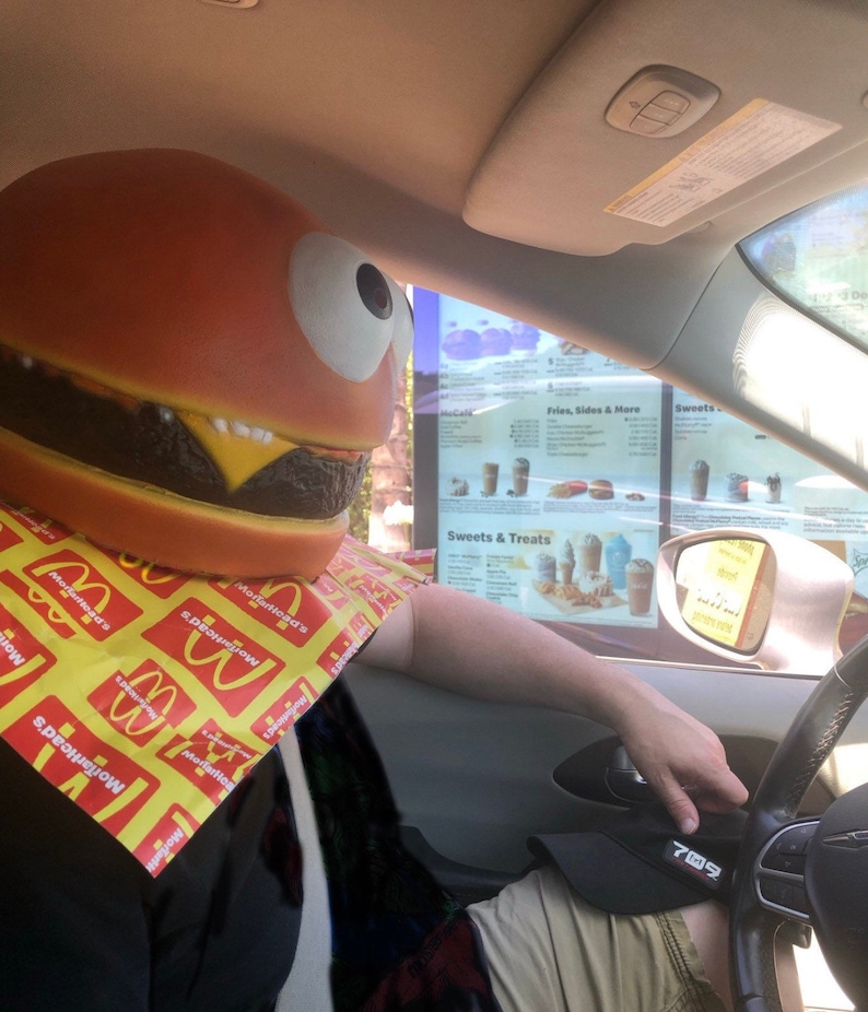 May include: A person wearing a large, red and brown hamburger costume with a yellow McDonald's napkin around their neck, sitting in a car at a drive-thru window. The menu board for the drive-thru is visible in the background.