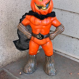 May include: A standing figurine of a pumpkin-headed character with a muscular orange body, wearing a black mesh cape, a dark belt with a small pumpkin buckle, and brown textured boots. The figure has a carved jack-o'-lantern face with a wide grin.