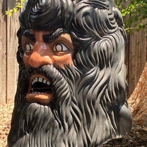 May include: A large, sculpted head with an angry expression. The face is tan with a dark beard and hair. The mouth is open, revealing teeth. The sculpture is likely made of a durable material and is mounted on a blue base.