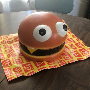 May include: A large, brown, foam hamburger with googly eyes and a yellow cheese slice. The hamburger is sitting on a yellow and red McDonald's themed paper.
