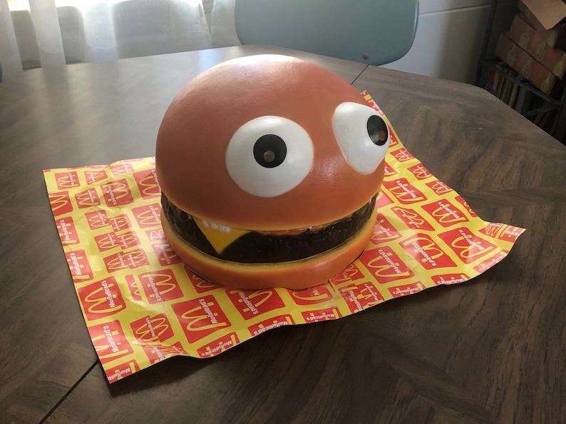May include: A large, brown, foam hamburger with googly eyes and a yellow cheese slice. The hamburger is sitting on a yellow and red McDonald's themed paper.
