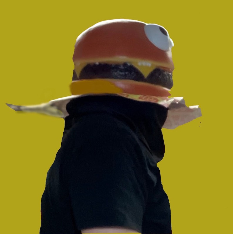 May include: A person wearing a brown and yellow hamburger costume with a white eye on the side. The costume is made of foam and has a realistic look.
