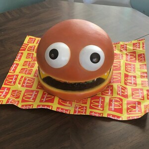 May include: A large, orange, plastic toy hamburger with googly eyes and a toothy grin. The toy is sitting on a yellow and red McDonald's themed paper.