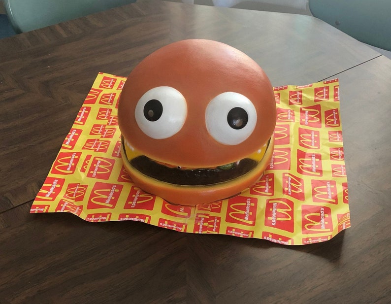 May include: A large, orange, plastic toy hamburger with googly eyes and a toothy grin. The toy is sitting on a yellow and red McDonald's themed paper.
