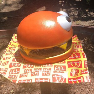 May include: A large orange and brown hamburger with a white googly eye, sitting on a yellow and red paper with the McDonald's logo repeated.