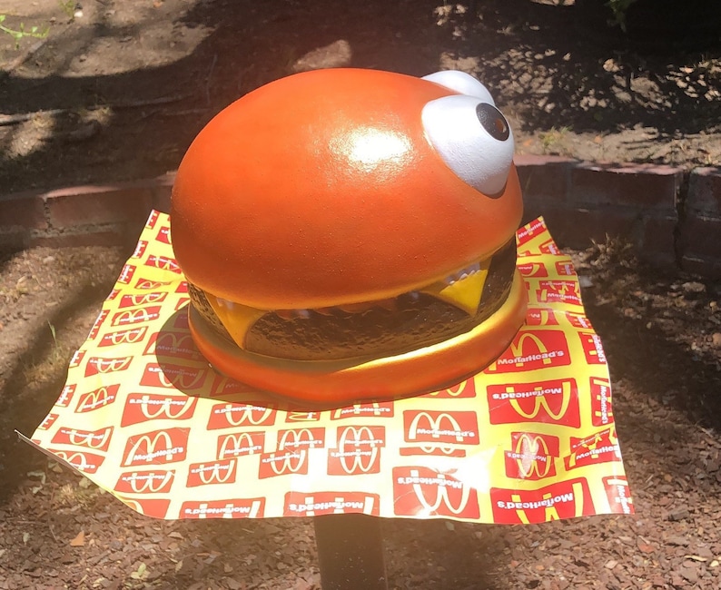May include: A large orange and brown hamburger with a white googly eye, sitting on a yellow and red paper with the McDonald's logo repeated.