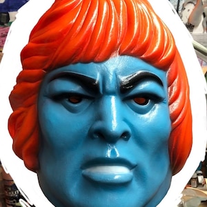 May include: A molded plastic mask featuring a bright orange, textured hairstyle and a solid blue face. The mask displays a stern facial expression with defined black eyebrows, deep-set eye openings, a prominent nose, and a closed, straight mouth.