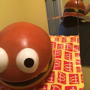 May include: A large, orange, round, hamburger-shaped toy with white eyes and a black mouth. The toy is sitting on a yellow and red patterned surface with the McDonald's logo repeated.