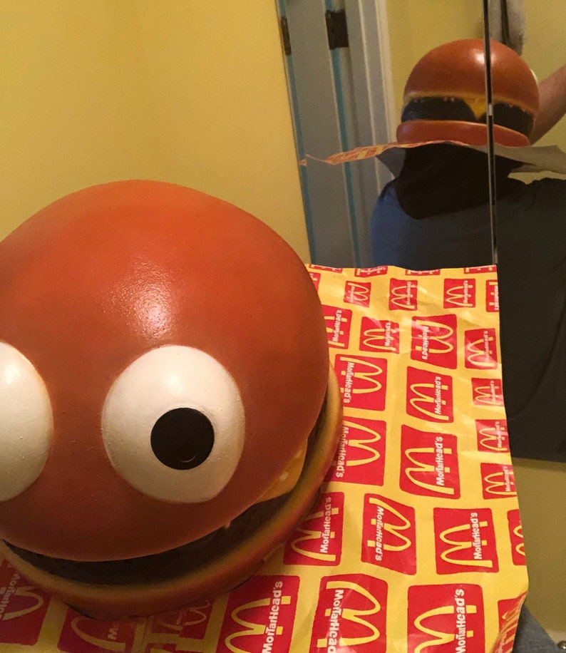 May include: A large, orange, round, hamburger-shaped toy with white eyes and a black mouth. The toy is sitting on a yellow and red patterned surface with the McDonald's logo repeated.