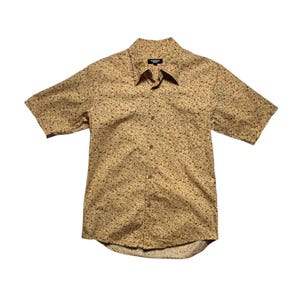 Può includere: Una camicia a maniche corte con bottoni e fantasia floreale. La camicia è di colore beige chiaro con piccoli dettagli floreali rossi e marroni. Il colletto è a punta e la camicia ha una tasca sul petto.