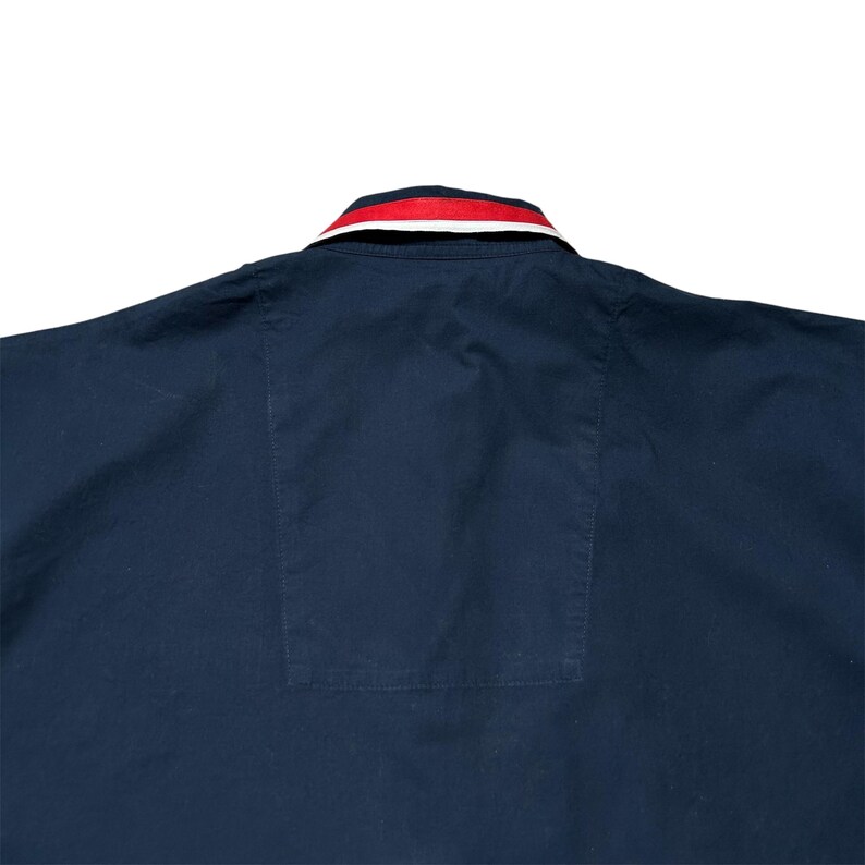 K&ouml;nnte beinhalten: Marineblaues Hemd mit rot-wei&szlig; gestreiftem Kragen. Das Hemd hat eine Tasche auf der unteren R&uuml;ckseite. Der Kragen hat ein klassisches Design, und das Hemd scheint aus einem glatten Stoff zu sein.