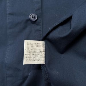 K&ouml;nnte beinhalten: Nahaufnahme eines marineblauen Hemdes mit Kn&ouml;pfen und einem angebrachten Etikett. Das Etikett enth&auml;lt japanischen Text. Der Stoff des Hemdes ist sichtbar, und der Knopf ist dunkel. Das Hemd besteht aus einem glatten Stoff.