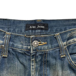 May include: Close-up of blue denim jeans with the brand name "Acne Jeans" on a black label. The jeans have a button closure and visible stitching details. The denim has a faded, worn appearance.