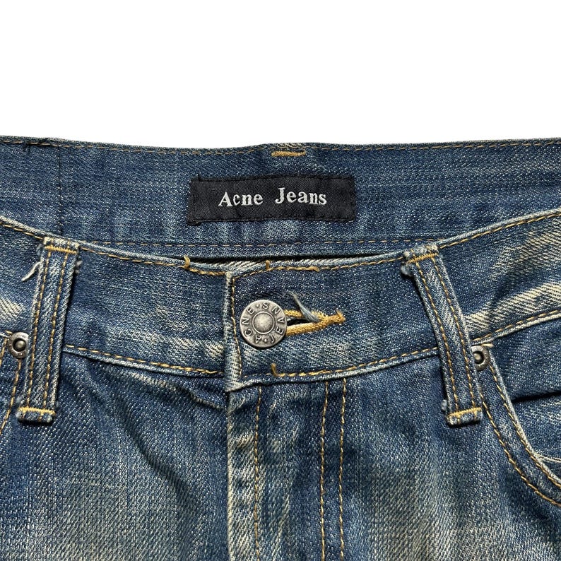 May include: Close-up of blue denim jeans with the brand name "Acne Jeans" on a black label. The jeans have a button closure and visible stitching details. The denim has a faded, worn appearance.