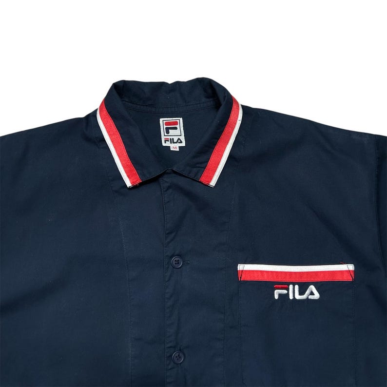 K&ouml;nnte beinhalten: Marineblaues Hemd mit rot-wei&szlig; gestreiftem Kragen und Taschenbesatz. Das FILA-Logo ist auf der Tasche und auf einem Etikett am Kragen aufgestickt. Das Hemd hat ein klassisches, sportliches Design f&uuml;r Freizeitkleidung.