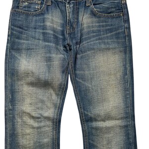 May include: Close-up of blue denim jeans. The jeans have a classic five-pocket design, with a button and zipper closure. The denim has a faded, worn look, with lighter patches and subtle distressing.