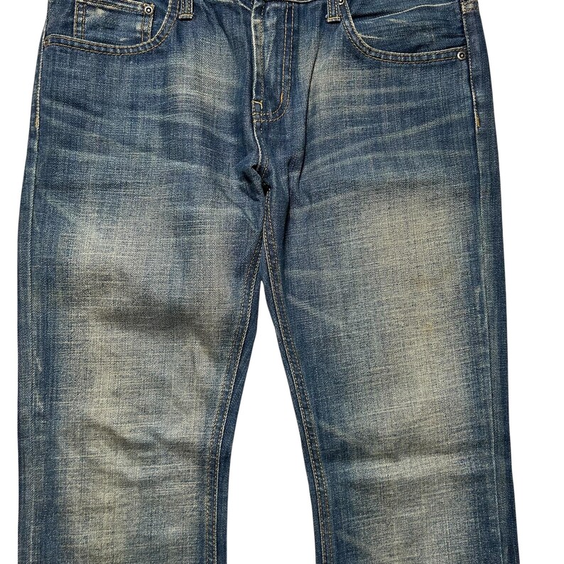 May include: Close-up of blue denim jeans. The jeans have a classic five-pocket design, with a button and zipper closure. The denim has a faded, worn look, with lighter patches and subtle distressing.