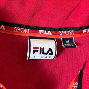 K&ouml;nnte beinhalten: Nahaufnahme eines roten Fila Sport-Hemdkragens. Der Kragen hat schwarze und orangefarbene Verzierungen mit dem Aufdruck "SPORT". Ein wei&szlig;es Etikett mit dem Fila-Logo und dem Wort "SPORT" ist sichtbar. Das Hemd ist Gr&ouml;&szlig;e M.