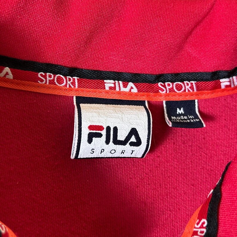 K&ouml;nnte beinhalten: Nahaufnahme eines roten Fila Sport-Hemdkragens. Der Kragen hat schwarze und orangefarbene Verzierungen mit dem Aufdruck "SPORT". Ein wei&szlig;es Etikett mit dem Fila-Logo und dem Wort "SPORT" ist sichtbar. Das Hemd ist Gr&ouml;&szlig;e M.