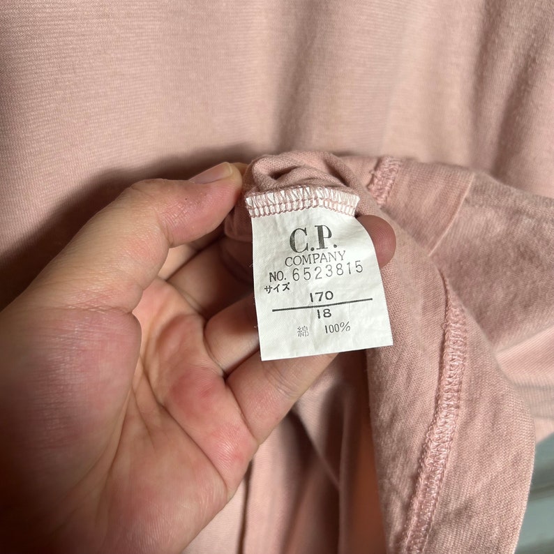 May include: A pink cotton t-shirt with a white tag that reads "C.P. Company, No. 6523815, サイズ 170, 18, 100%"