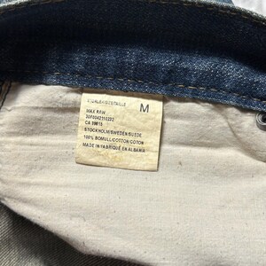 May include: Close-up of a denim garment's interior, revealing a tag with text. The tag includes the size "M", product details, and the text "STOCKHOLM/SWEDEN/SUEDE" and "MADE IN/FABRIQU&Eacute; EN ALBANIA".
