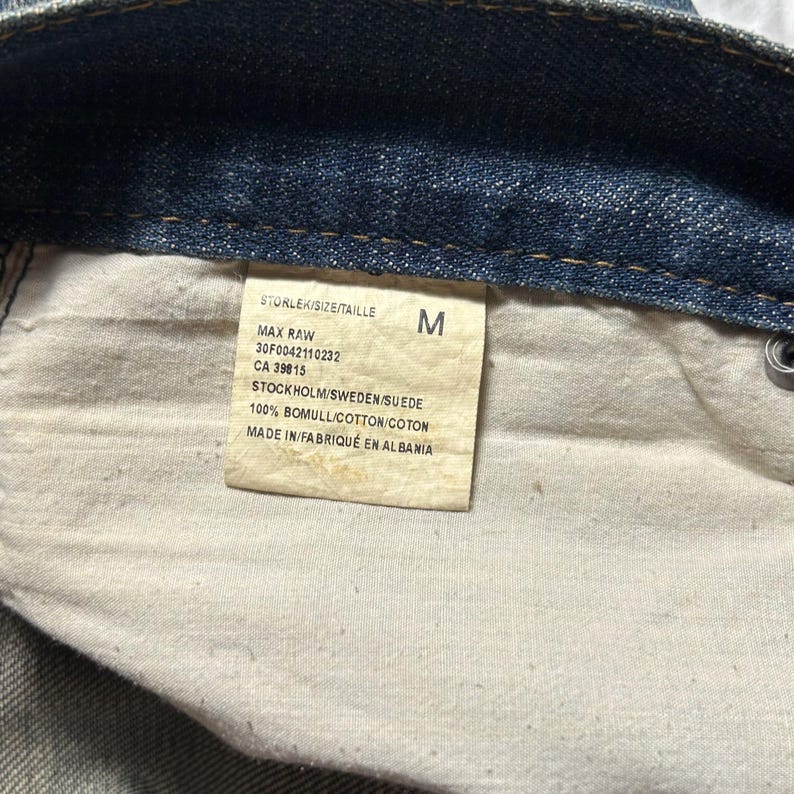 May include: Close-up of a denim garment's interior, revealing a tag with text. The tag includes the size "M", product details, and the text "STOCKHOLM/SWEDEN/SUEDE" and "MADE IN/FABRIQU&Eacute; EN ALBANIA".