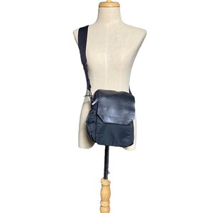 Könnte beinhalten: Eine schwarze Umhängetasche aus Nylon mit einem Lederklappenverschluss und einem Reißverschluss. Die Tasche hat einen verstellbaren Schultergurt für optimalen Tragekomfort.