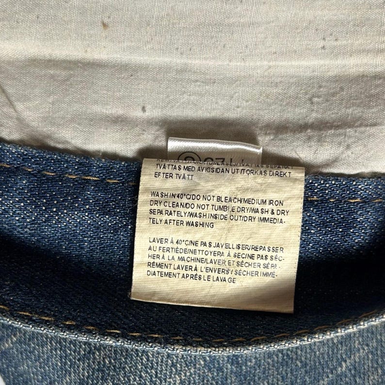 May include: Close-up of a denim garment label with care instructions. The label is rectangular, with text in multiple languages, including English and French. The denim fabric is a medium blue color.