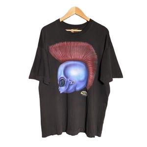 May include: Black t-shirt with a graphic print of a blue alien head with a red mohawk. The alien has one large eye and a small mouth. The text "TRIBE" is printed on the bottom right of the graphic.