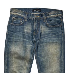 May include: Blue denim jeans with a faded wash. The jeans have a classic five-pocket design, a button and zipper closure, and the brand name "Acne Jeans" on a label at the waistband. The denim has a worn appearance.