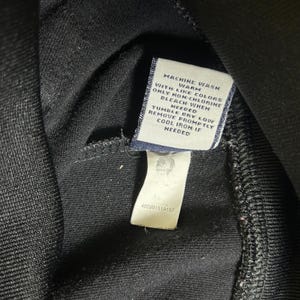 K&ouml;nnte beinhalten: Nahaufnahme eines schwarzen Kleidungsst&uuml;cks mit einem Pflegeetikett. Das Etikett ist wei&szlig; mit schwarzem Text, einschlie&szlig;lich Waschanweisungen und dem Text "MACHINE WASH WARM WITH LIKE COLORS ONLY NON-CHLORINE BLEACH WHEN NEEDED".