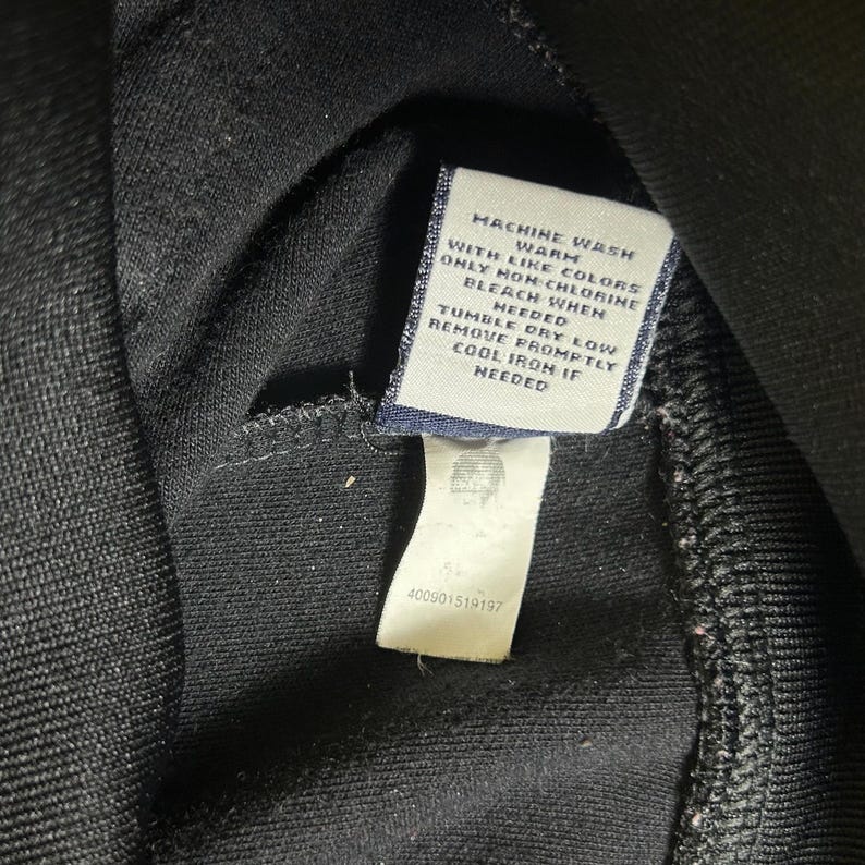 K&ouml;nnte beinhalten: Nahaufnahme eines schwarzen Kleidungsst&uuml;cks mit einem Pflegeetikett. Das Etikett ist wei&szlig; mit schwarzem Text, einschlie&szlig;lich Waschanweisungen und dem Text "MACHINE WASH WARM WITH LIKE COLORS ONLY NON-CHLORINE BLEACH WHEN NEEDED".