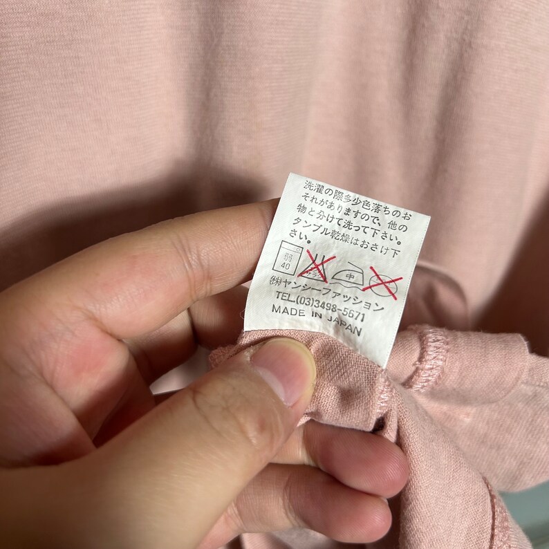 May include: A pink cotton t-shirt with a white care tag attached. The tag has instructions for washing and drying the shirt. The tag also includes the brand name "Yansey Fashion" and the phone number (03)3498-5671. The tag states "Made in Japan."