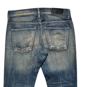 May include: Close-up of the back of blue denim jeans. The jeans have two back pockets and a leather patch at the waistband. The denim has a faded, worn look with subtle distressing.