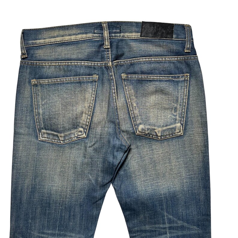 May include: Close-up of the back of blue denim jeans. The jeans have two back pockets and a leather patch at the waistband. The denim has a faded, worn look with subtle distressing.