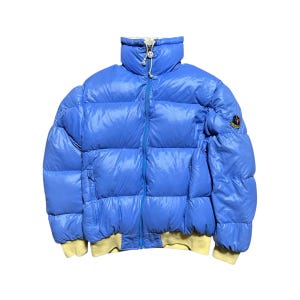 Puede incluir: Chaqueta acolchada azul claro con cuello alto y cierre de cremallera. La chaqueta tiene forro amarillo en los puños y el dobladillo. Un pequeño parche con el logotipo es visible en la manga superior izquierda. La chaqueta está diseñada para el frío.