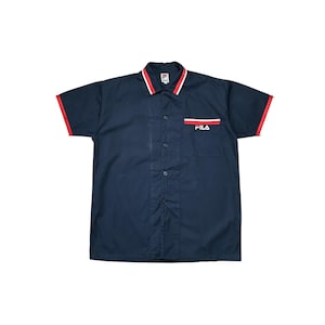 K&ouml;nnte beinhalten: Marineblaues Kurzarmhemd mit roter Paspelierung an &Auml;rmeln und Kragen. Der Kragen hat einen wei&szlig;en und roten Streifen. Das Hemd hat eine Tasche mit dem Wort "FILA" in Rot und Wei&szlig;.