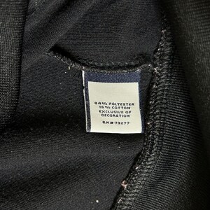 K&ouml;nnte beinhalten: Nahaufnahme eines schwarzen Kleidungsst&uuml;cks mit einem Etikett. Das Etikett ist wei&szlig; mit marineblauem Text, der lautet: "84% Polyester 16% Baumwolle Exclusive of Decoration RN #73277". Das Kleidungsst&uuml;ck besteht aus einem weichen, strukturierten Stoff.