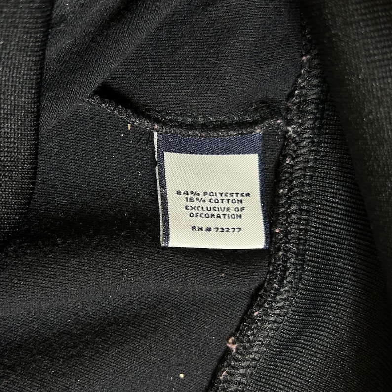 K&ouml;nnte beinhalten: Nahaufnahme eines schwarzen Kleidungsst&uuml;cks mit einem Etikett. Das Etikett ist wei&szlig; mit marineblauem Text, der lautet: "84% Polyester 16% Baumwolle Exclusive of Decoration RN #73277". Das Kleidungsst&uuml;ck besteht aus einem weichen, strukturierten Stoff.