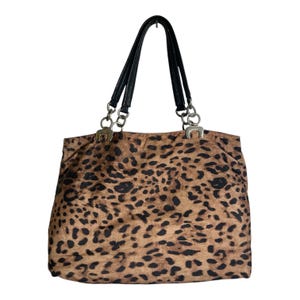 May include: A tote bag featuring a tan and black leopard print pattern. The bag has two black handles attached to the main body with silver-toned metal chain links and rectangular hardware connectors at the top corners of the bag's opening.
