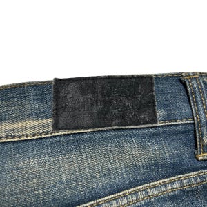 May include: Close-up of blue denim jeans with a black leather patch. The patch is embossed with the brand name "Acne". The jeans have visible stitching and a faded wash.