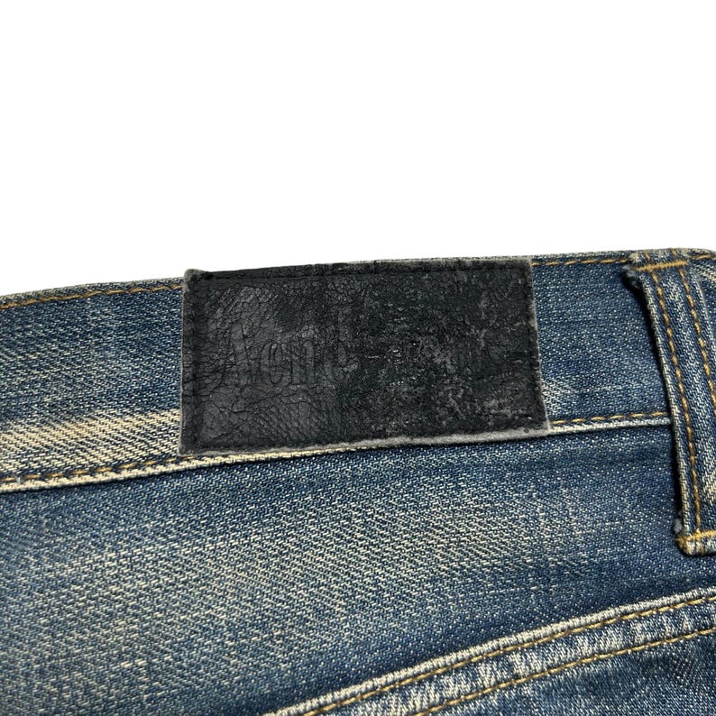 May include: Close-up of blue denim jeans with a black leather patch. The patch is embossed with the brand name "Acne". The jeans have visible stitching and a faded wash.