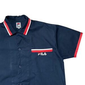 K&ouml;nnte beinhalten: Marineblaues Kurzarmhemd mit rot-wei&szlig;em Besatz an Kragen, Tasche und &Auml;rmeln. Das Fila-Logo ist auf der Tasche und dem Kragen zu sehen. Das Hemd hat eine Knopfleiste.