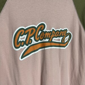 May include: A beige and green baseball-style t-shirt with the text "C.P. Company" in a brown and white graphic.