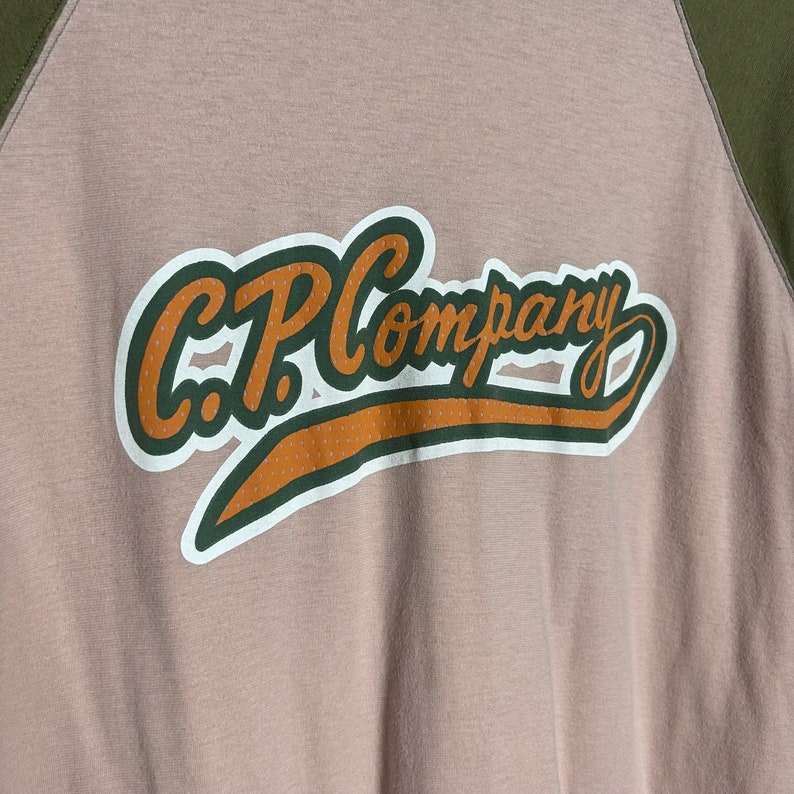 May include: A beige and green baseball-style t-shirt with the text "C.P. Company" in a brown and white graphic.