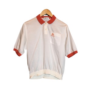 May include: White polo shirt with a red trim around the collar and sleeves. The shirt has a small embroidered logo on the left chest that says "Burberry".