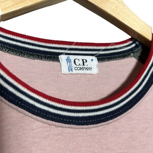 May include: Pink t-shirt with a black, white, and red striped collar. The shirt has a label that reads "C.P. COMPANY".
