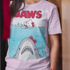 May include: Light pink t-shirt featuring a cartoon graphic of a shark with a red bow and a cartoon cat in the water. The word "JAWS" is printed in red above the graphic. The shirt is short-sleeved.