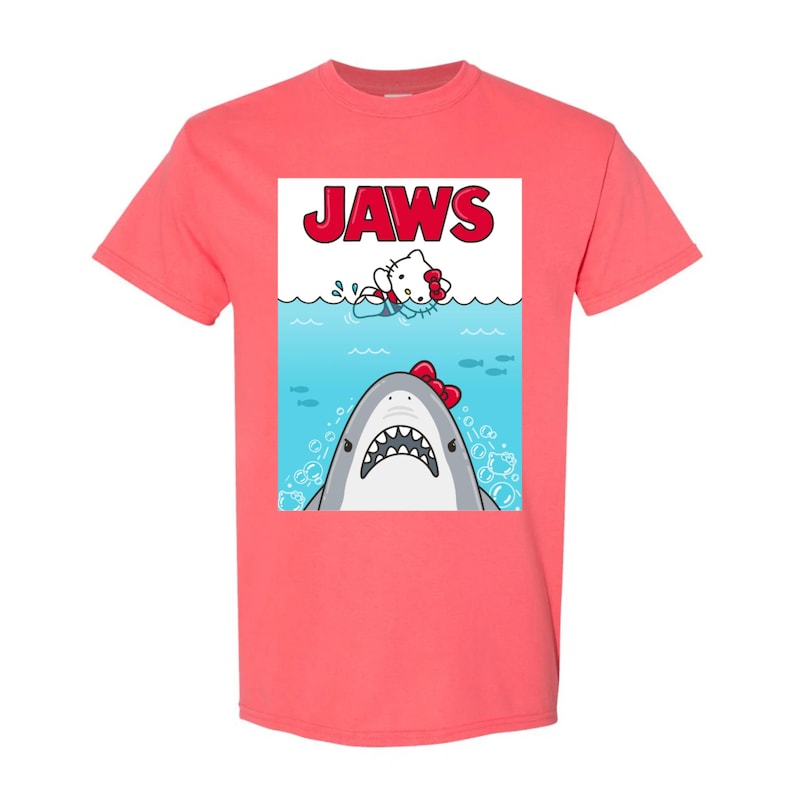 May include: Coral-colored t-shirt featuring a graphic of a shark with a red bow and a cartoon cat in the water, with the word "JAWS" in red letters above. The design is a parody of the movie poster.